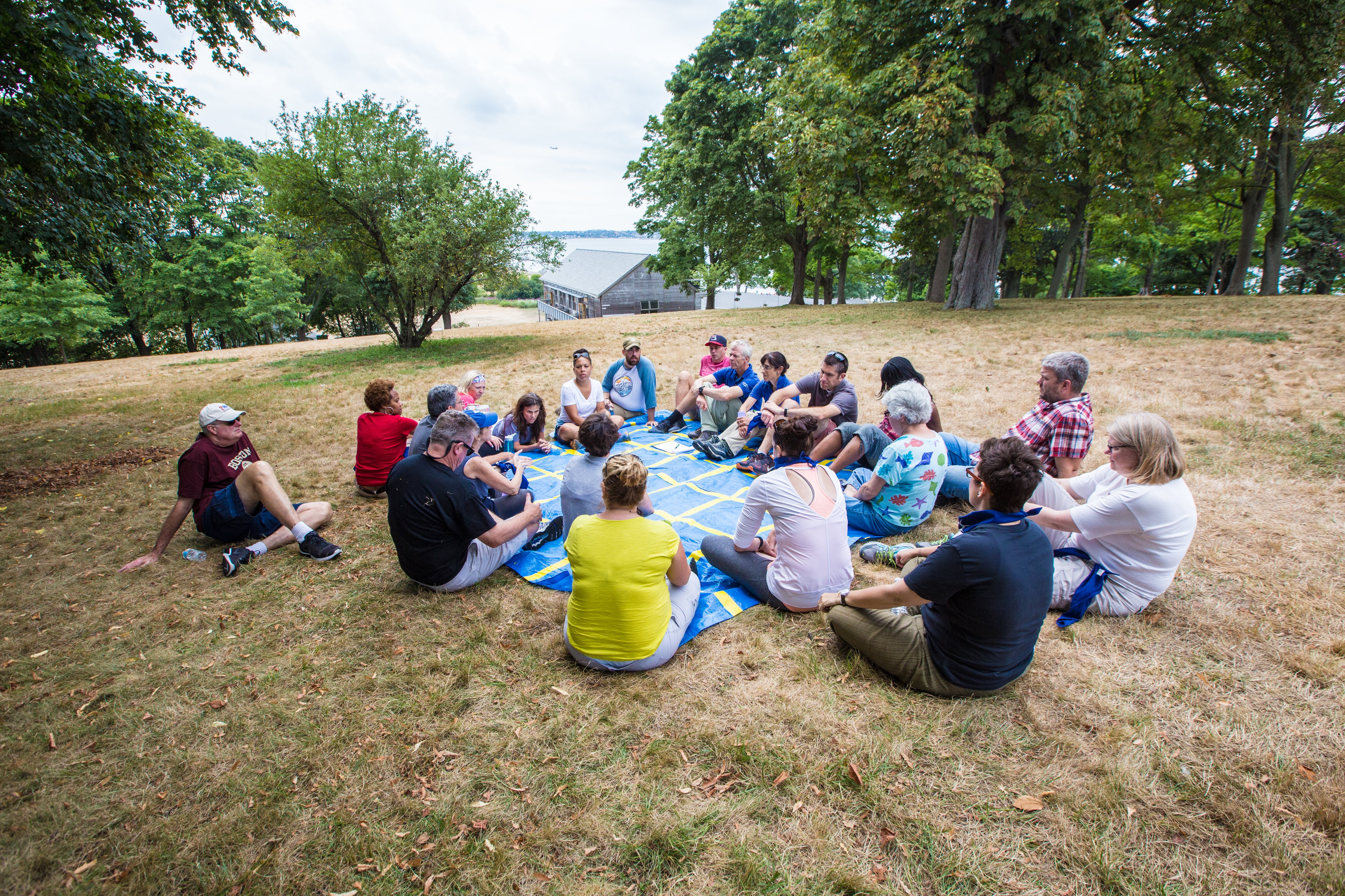 The image shows a group of people sitting in a circle on a blue mat in a grassy area. They appear to be engaged in a discussion or activity. Trees surround the group, and a body of water is visible in the background. The scene suggests an outdoor gathering or meeting.